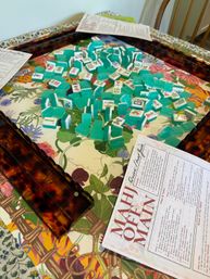 Scattered turquoise-green mahjong tiles and amber tile racks on a floral tabletop with printed mahjong rules, ready for a casual indoor game