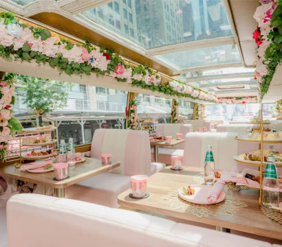 Glass-roofed city tour bus interior decorated with pink booths and lush floral garlands, tables set for afternoon tea with tiered pastry stands, plates, napkins and sparkling water.