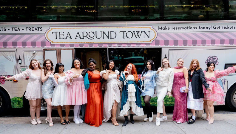 Smiling group of women in colorful vintage and modern dresses posing together in front of a pink tea-tour bus parked on a city sidewalk.