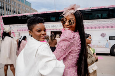 Two stylish women smiling at an outdoor tea event beside a pink floral tea bus on a city street