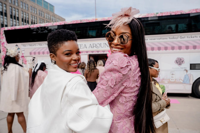 Two stylish women smiling at an outdoor tea event beside a pink floral tea bus on a city street