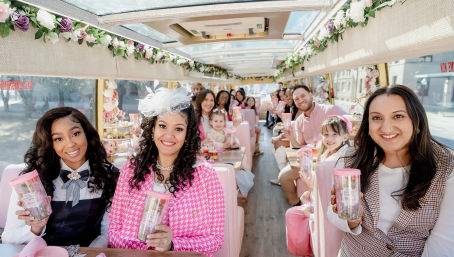 Smiling group of adults and children on a pink, flower‑decorated party bus/trolley holding matching pink tumblers during a daytime city celebration