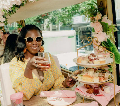 Smiling woman in a yellow dress and sunglasses toasting with a glass during a floral‑decorated afternoon tea on a city bus, with a tiered tray of sandwiches and pastries by the window.