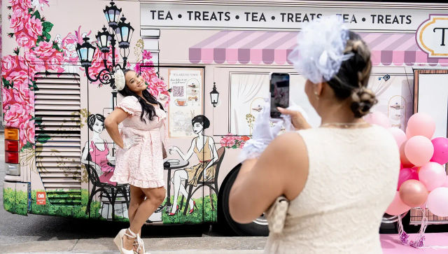 Smiling woman in a pink vintage dress poses in front of a floral pink "Tea • Treats" tea truck mural while another person photographs her with a smartphone next to pink balloons.