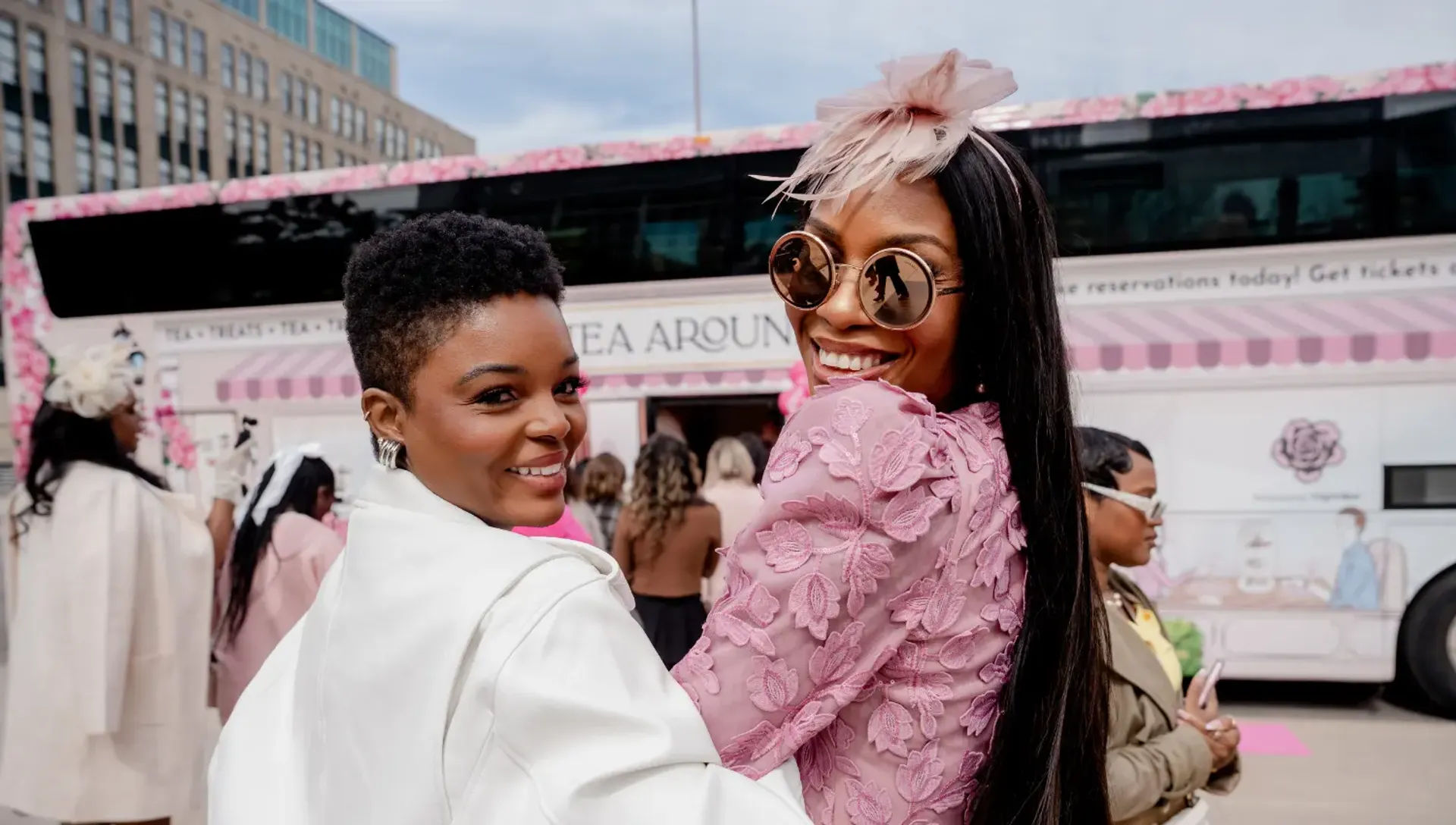 Two fashionable women smiling at a pink-themed outdoor tea pop-up in a city plaza — one in pink lace with round sunglasses and a fascinator, the other in a white coat.