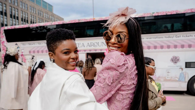 Two fashionable women smiling at a pink-themed outdoor tea pop-up in a city plaza — one in pink lace with round sunglasses and a fascinator, the other in a white coat.