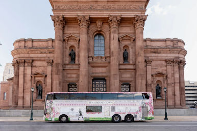 Floral-wrapped double-decker tour bus parked on a city street in front of a grand neoclassical sandstone building with tall Corinthian columns, arched windows and statue-filled niches.