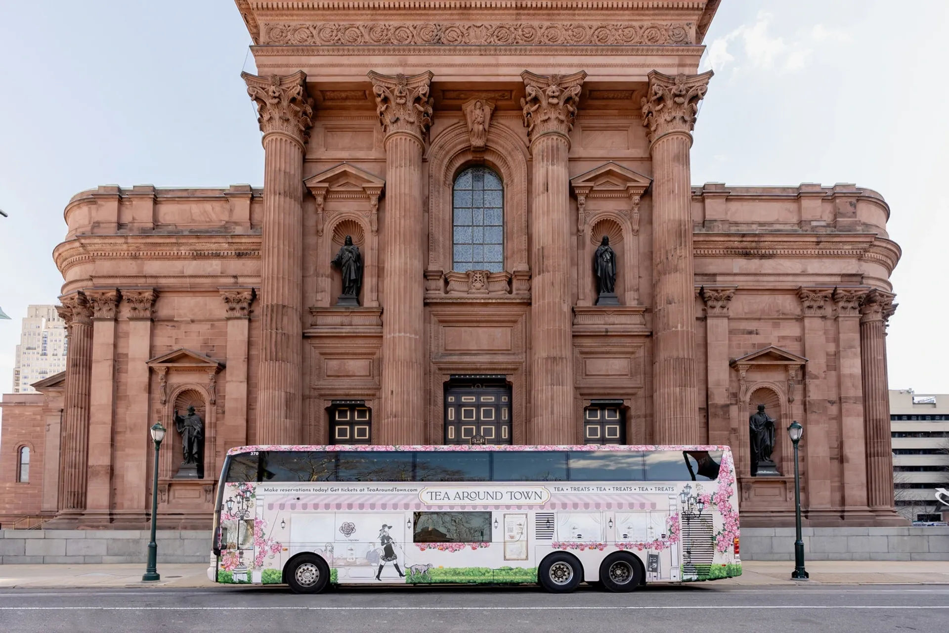 Floral-wrapped double-decker tour bus parked on a city street in front of a grand neoclassical sandstone building with tall Corinthian columns, arched windows and statue-filled niches.