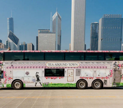 Pink floral tea-themed double-decker tour bus parked on a bridge with the Chicago skyline of modern skyscrapers under a clear blue sky