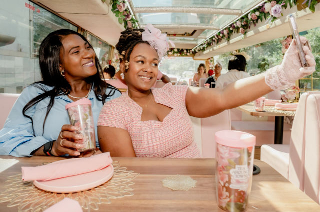 Two smiling women in pastel outfits take a selfie inside a sunlit, floral-decorated pink tea party bus, holding floral tumblers at a table set with pink plates and napkins.