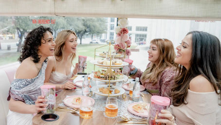 Four friends enjoying a pink-themed afternoon tea with pastries and drinks on a decorated party bus with city park views