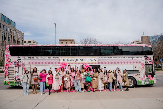 Group of women in pink and spring outfits posing with balloons in front of a floral-wrapped double-decker tea-themed bus on a downtown city street.