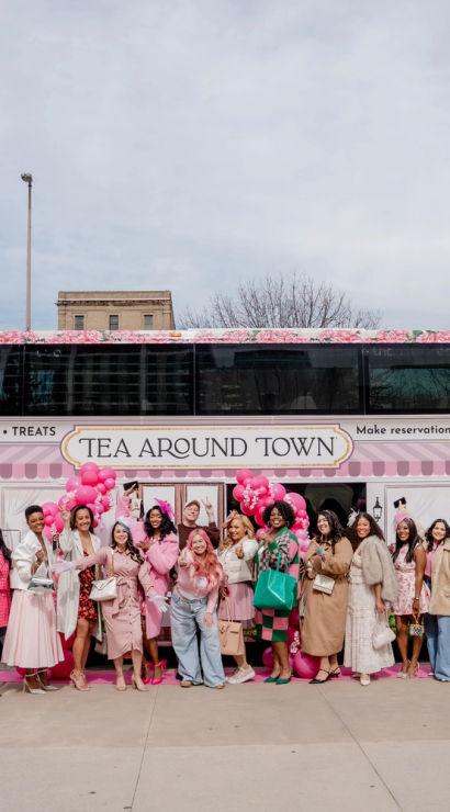 Group of women in pink and spring outfits posing with balloons in front of a floral-wrapped double-decker tea-themed bus on a downtown city street.
