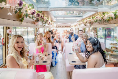 Smiling friends toasting on a flower-adorned pink party trolley in a sunny city street, enjoying a daytime bachelorette/brunch celebration with drinks and pastel decor.