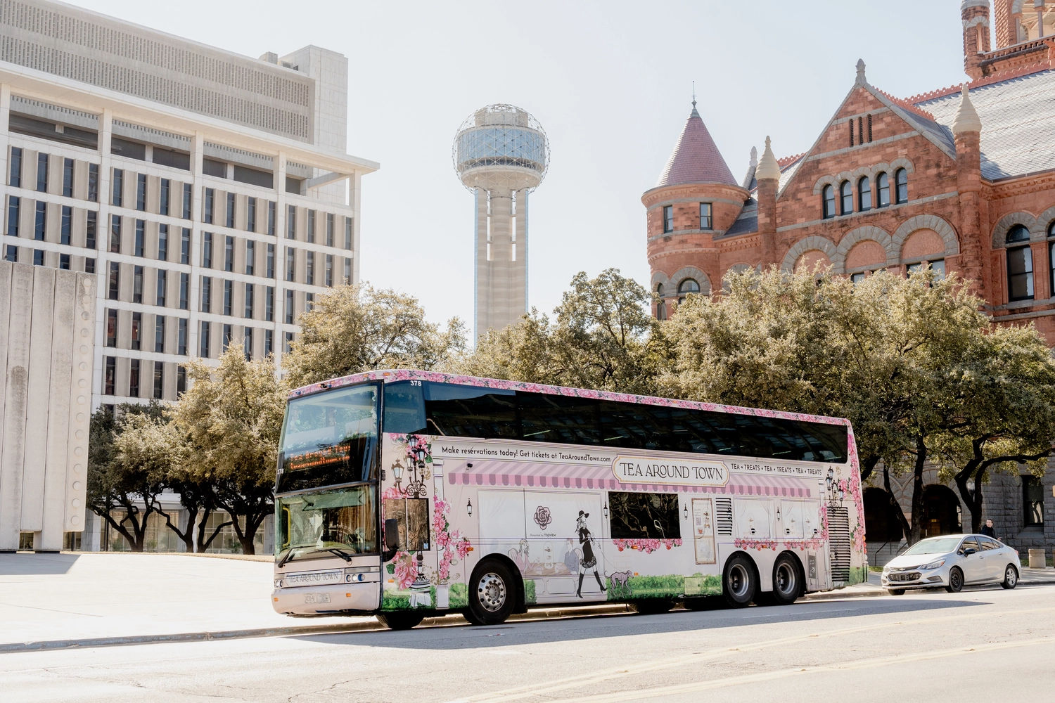 Cheerful pink floral double-decker tea-themed tour bus parked on a sunny street in downtown Dallas, with the Reunion Tower sphere and a historic red-brick Romanesque building framed by oak trees in the background.