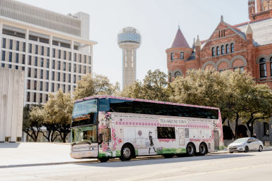 Cheerful pink floral double-decker tea-themed tour bus parked on a sunny street in downtown Dallas, with the Reunion Tower sphere and a historic red-brick Romanesque building framed by oak trees in the background.