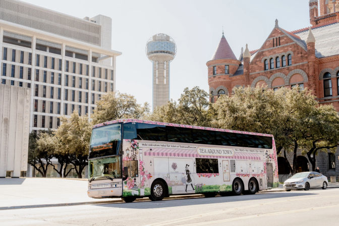 Cheerful pink floral double-decker tea-themed tour bus parked on a sunny street in downtown Dallas, with the Reunion Tower sphere and a historic red-brick Romanesque building framed by oak trees in the background.