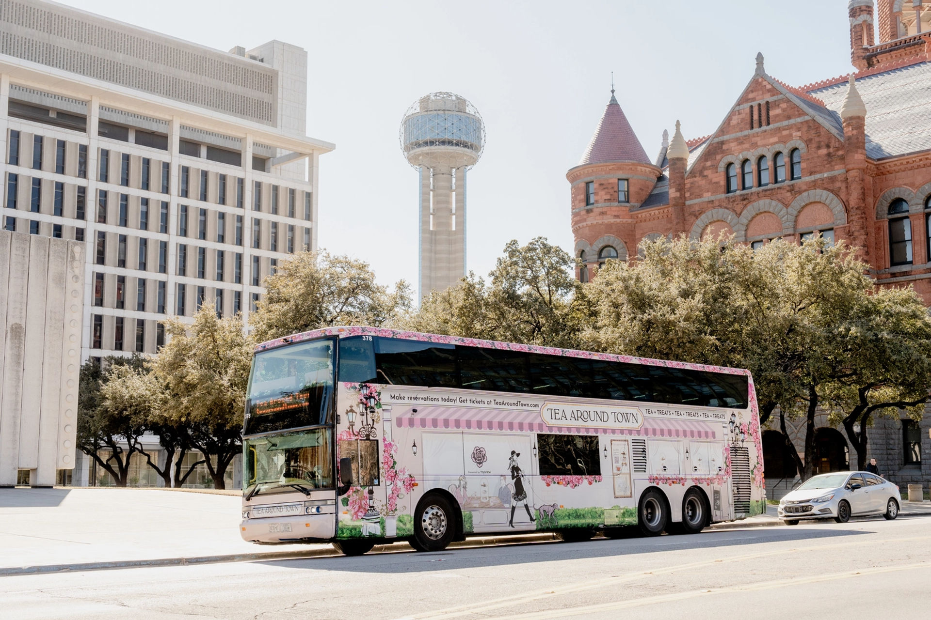 Cheerful pink floral double-decker tea-themed tour bus parked on a sunny street in downtown Dallas, with the Reunion Tower sphere and a historic red-brick Romanesque building framed by oak trees in the background.