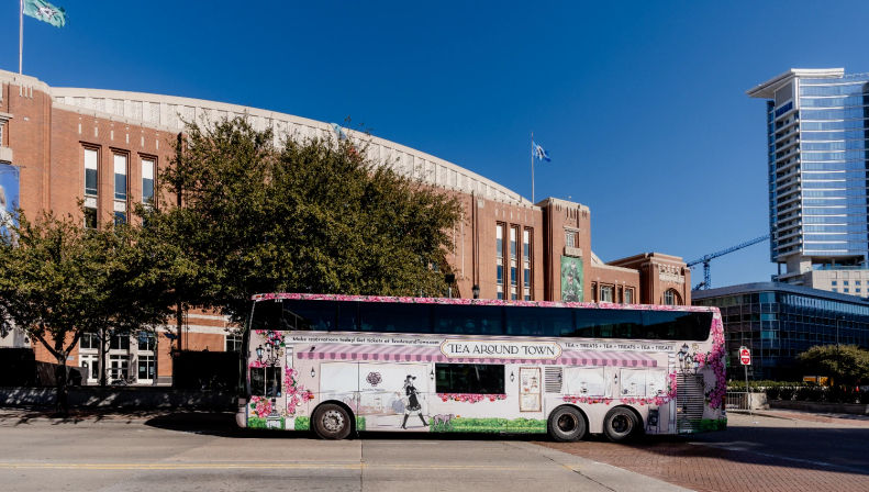 Cheery pink floral double-decker tea tour bus parked on a downtown street in front of a large brick arena and glass high-rise under a clear blue sky
