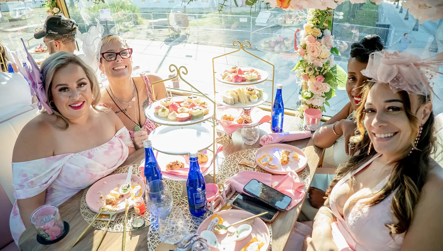 Four smiling women in pink dresses and fascinators enjoying a sunny waterfront afternoon tea with three-tier pastry stands, pink tableware, floral garland and blue water bottles.