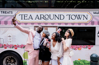 Four friends smiling and taking a selfie in front of a pink floral tea truck on a city street, each holding clear tea tumblers.