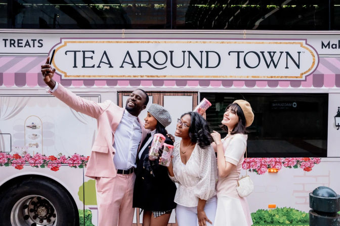 Four friends smiling and taking a selfie in front of a pink floral tea truck on a city street, each holding clear tea tumblers.