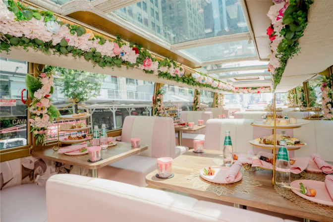 Pastel-pink, floral-decorated panoramic dining bus set for afternoon tea with tiered pastry trays, bottled water and city street views through skylights.