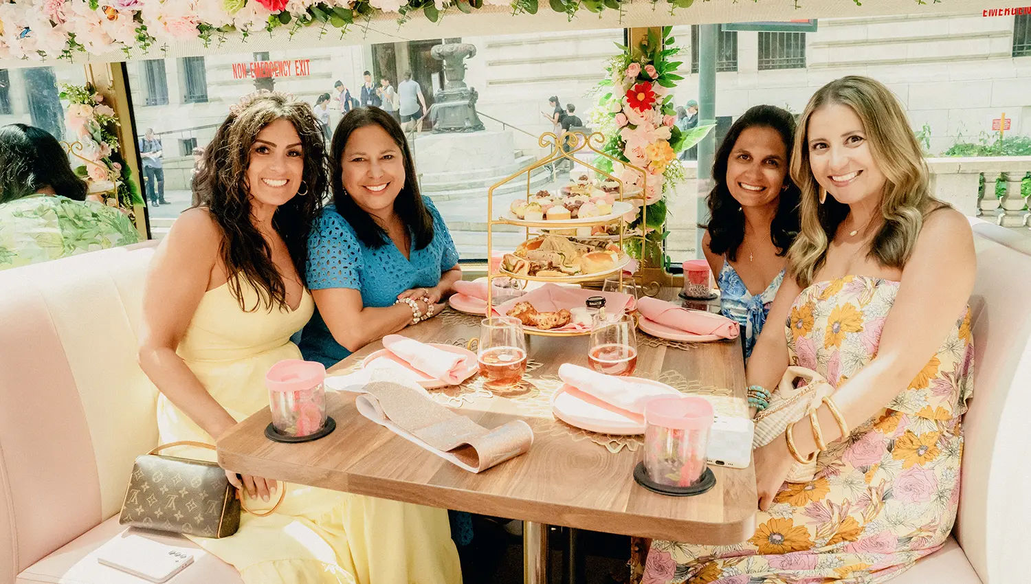 Four women in colorful summer dresses enjoying urban afternoon tea at a floral-decorated table with a tiered tray of pastries, rosé glasses, pink napkins and a city street view through the window.