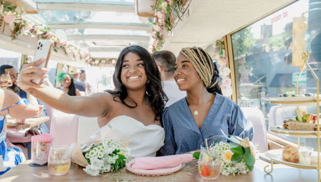 Two smiling women taking a selfie on a floral‑decorated party bus during a daytime bridal brunch, with bouquets, tea glasses and pastries on the table and city streets visible through the windows.