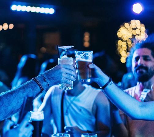 Friends clinking a tropical cocktail and a beer pint under blue club lighting, celebratory bar scene with bokeh string lights and lively nightlife vibe