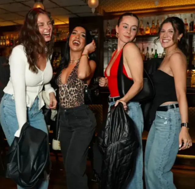 Four friends laughing on a night out in a lively bar, wearing jeans and trendy tops and holding jackets in front of illuminated liquor shelves — upbeat city nightlife vibe.