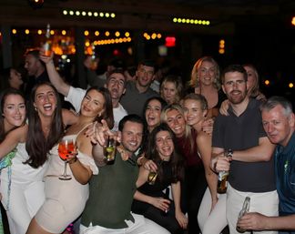 Lively nightlife scene: large group of friends posing and smiling inside a crowded bar, holding cocktails and beer bottles under warm string lights.