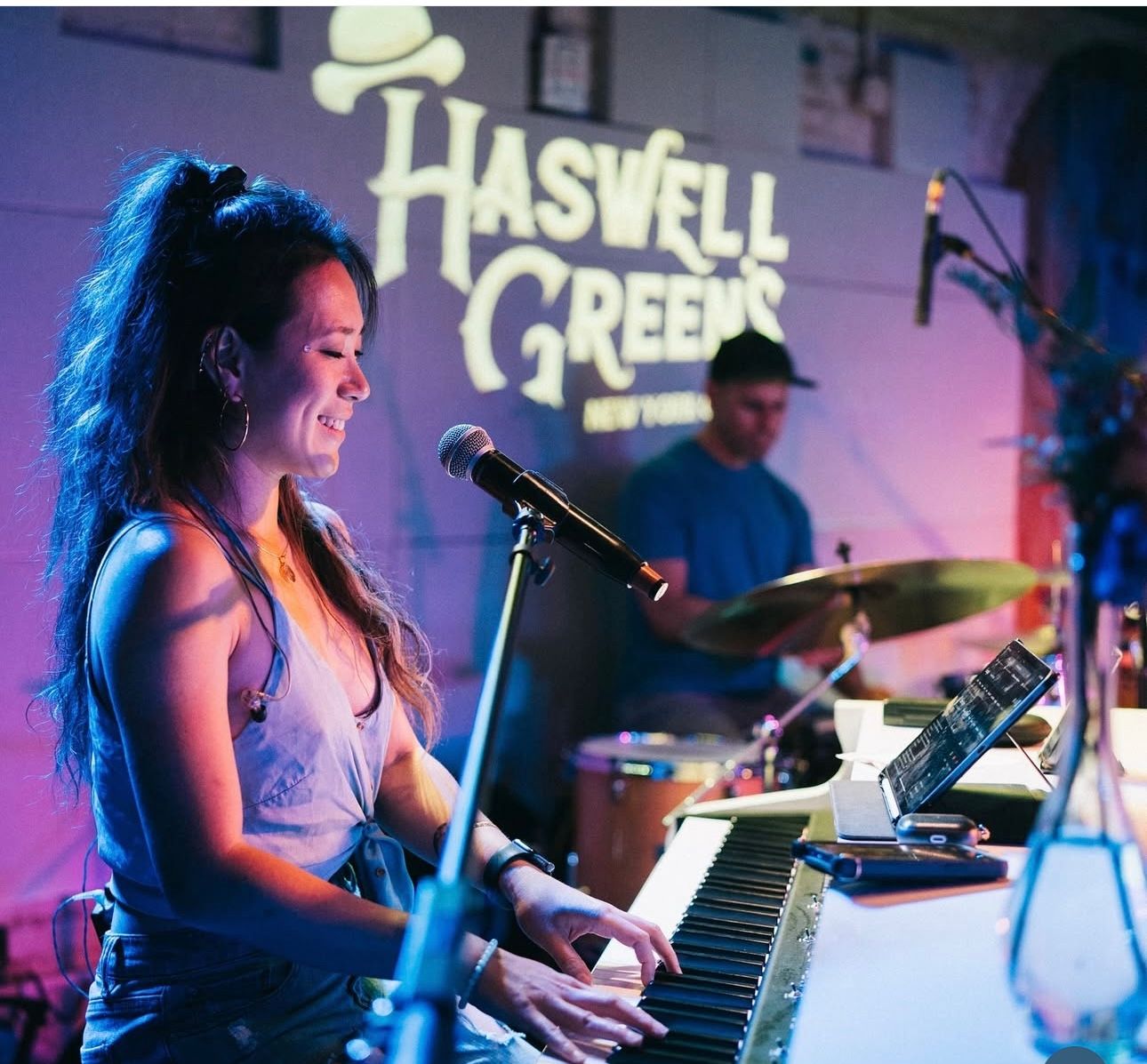 Smiling singer-keyboardist playing electric piano and singing into a microphone on a colorful-lit indoor stage, drummer in the background and venue signage projected on the wall.