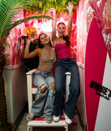 Two friends smiling and raising their arms in a vibrant tropical-themed photo booth with pink palm-print wallpaper, neon lighting, surfboard prop and lush greenery for a beachy party vibe.
