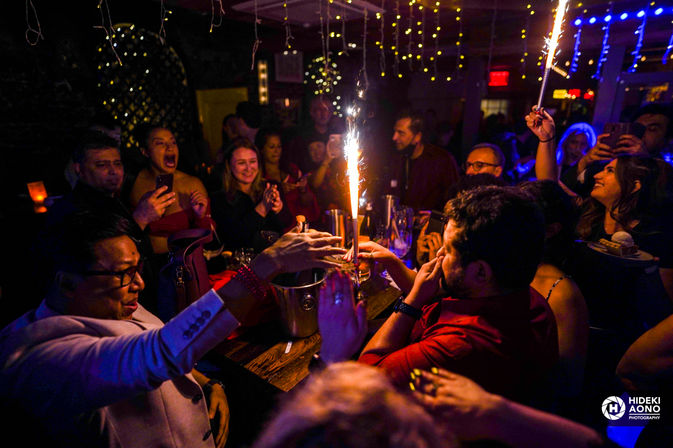 Lively bar party: crowd gathered around a table with champagne bottle sparklers, clinking glasses, cheering and taking photos under hanging fairy lights.