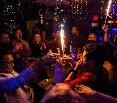 Lively bar party: crowd gathered around a table with champagne bottle sparklers, clinking glasses, cheering and taking photos under hanging fairy lights.
