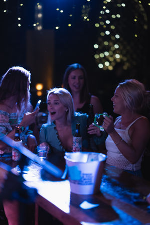 Group of friends laughing at a lively outdoor bar at night, holding beers, a tambourine and a phone beneath warm bokeh string lights