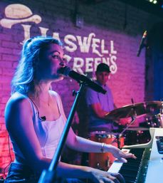 Female singer playing keyboard and singing into a microphone, accompanied by a drummer under purple-blue stage lighting in an intimate brick-walled live music venue.