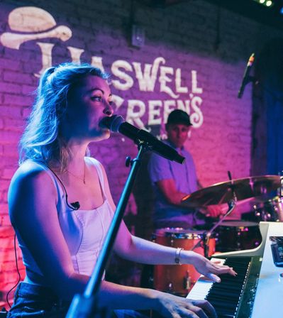 Female singer playing keyboard and singing into a microphone, accompanied by a drummer under purple-blue stage lighting in an intimate brick-walled live music venue.