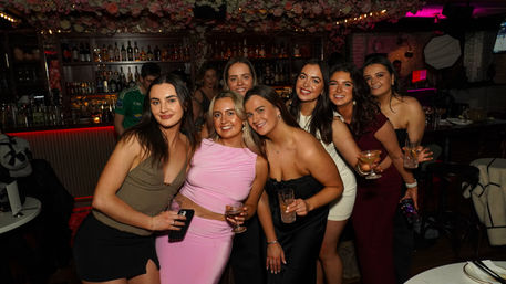 Smiling group of women in stylish party outfits holding cocktails and posing together at a dimly lit bar with floral ceiling decor and shelves of bottles
