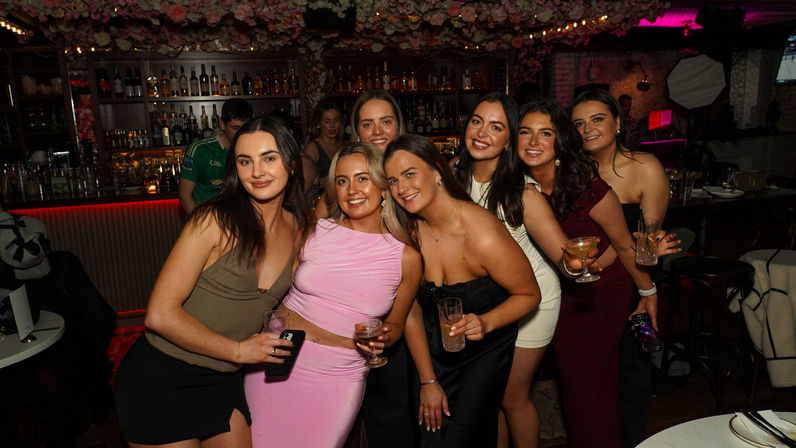 Smiling group of women in stylish party outfits holding cocktails and posing together at a dimly lit bar with floral ceiling decor and shelves of bottles