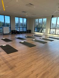 Sunlit fitness studio with wood floors and floor-to-ceiling windows showing palm trees, black exercise mats spaced evenly across the room with folded towels ready for class.