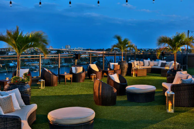 Cozy rooftop lounge at dusk — wicker sofas and ottomans on green turf, palm trees, string lights and lanterns overlooking a waterfront marina and city skyline.