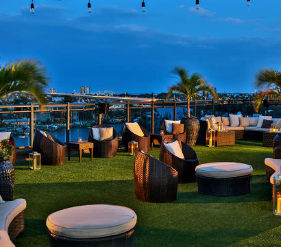Cozy rooftop lounge at dusk — wicker sofas and ottomans on green turf, palm trees, string lights and lanterns overlooking a waterfront marina and city skyline.