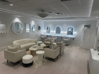 Bright modern beauty salon lounge with cream sofa and armchairs around coffee tables, light wood floors, decorative round wall mirrors, LED-lit arched vanity mirrors with white styling stools and a central chandelier.