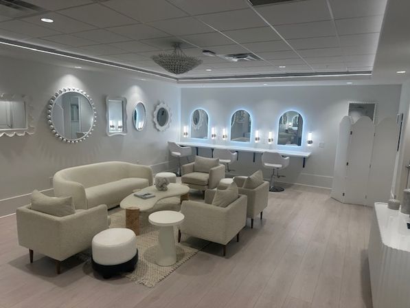 Bright modern beauty salon lounge with cream sofa and armchairs around coffee tables, light wood floors, decorative round wall mirrors, LED-lit arched vanity mirrors with white styling stools and a central chandelier.