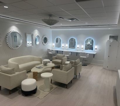 Bright modern beauty salon lounge with cream sofa and armchairs around coffee tables, light wood floors, decorative round wall mirrors, LED-lit arched vanity mirrors with white styling stools and a central chandelier.