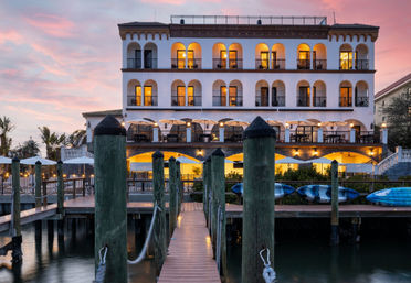 Sunset-lit Mediterranean-style waterfront hotel with arched balconies, outdoor dining terrace, wooden pier leading from a marina and kayaks on the shore.
