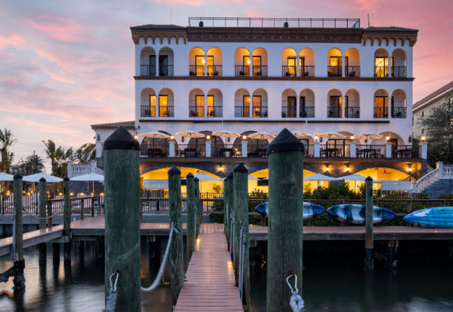 Sunset-lit Mediterranean-style waterfront hotel with arched balconies, outdoor dining terrace, wooden pier leading from a marina and kayaks on the shore.