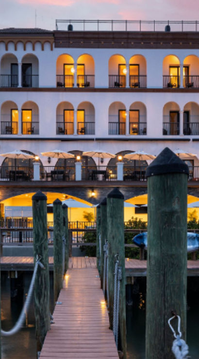 Sunset-lit Mediterranean-style waterfront hotel with arched balconies, outdoor dining terrace, wooden pier leading from a marina and kayaks on the shore.